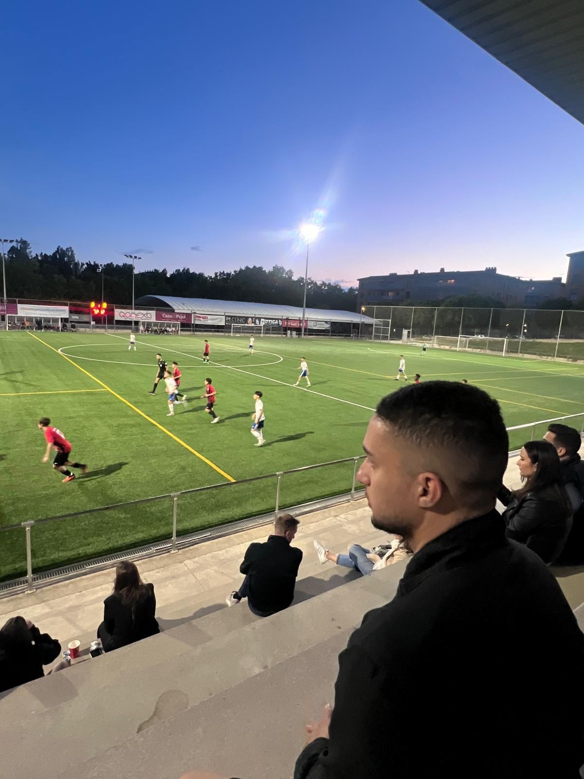 People watching a soccer match in an outdoor stadium during sunset with players on the field.